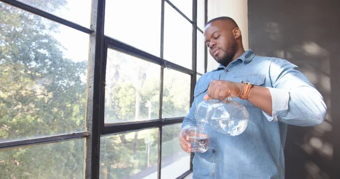 African American man enjoying fresh water from glass pitcher by window