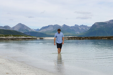 Young man walking barefoot in the crystal clear water of Sommaroy beach in Norway. Traveler enjoying summer vacation on the arctic coast of northern Norway. © vaitekune