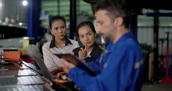 Male EV technician reviews tablet near exposed battery components while two Asian female customers observe and discuss inspection details during electric vehicle consultation in modern workshop.