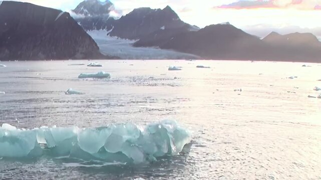 A blue iceberg floats on cold water beneath rugged mountain slopes. Secondary visible features include an icy bay, with natural contrast and texture defining the frame.