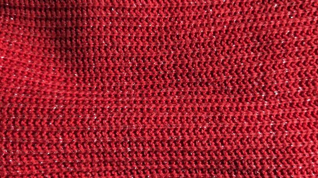 Close up of red knitted sweater fabric texture with shiny metallic glitter thread, smoothly panning right to showcase festive winter clothing, Christmas fashion, and warm cozy wool material background