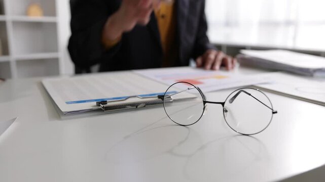 Businessman analyzing financial documents and signing a contract. Close-up on glasses with a businessman in the background analyzing financial statistics and signing important paperwork in the office.