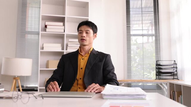 Exhausted young Asian manager finishing work, closing laptop, and stretching his neck. Man feeling tired from overworking in the office.