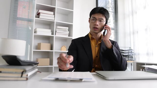 Young Asian professional man wearing glasses talking on a mobile phone and taking notes on a clipboard document while working at his desk.