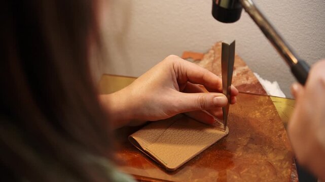 Female artisan punches stitching holes in a leather wallet using a pricking iron and hammer. Handmade leathercraft process for fashion accessories, design and small business