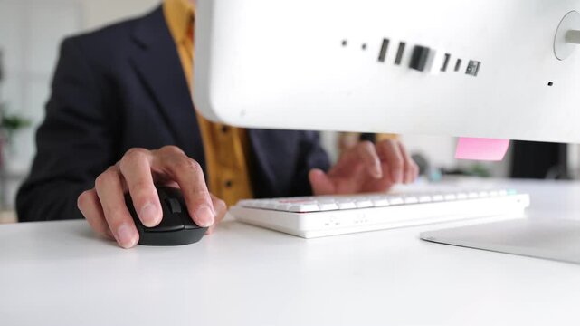 Businessman working on computer using mouse and typing on keyboard. His hands are clicking the mouse and typing on a modern white computer keyboard.