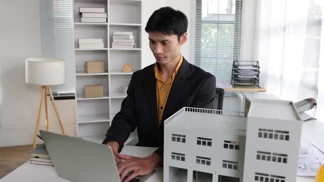Professional Asian architect typing on a laptop and checking a building model. Concentrated man working on a construction project in his office.