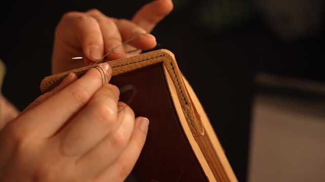 Female artisan hand stitches a leather accessory with green thread using needles. Traditional leathercraft process for handmade goods, fashion and small business