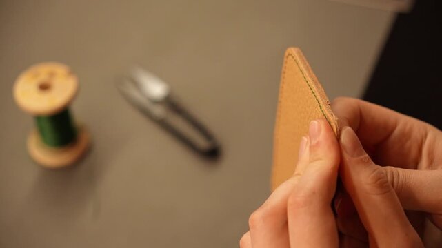 Female artisan hand stitches a leather accessory with green thread using needles. Traditional leathercraft process for handmade goods, fashion and small business