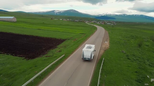 A white camper rides on an empty asphalt road through a vast green landscape with snow-capped mountains in the background, embodying the concept of freedom, travel and adventure vacation in Georgia