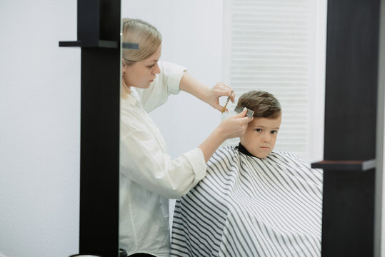 A photo from the life of a hairdresser's client, a dark-haired boy. A female hairdresser at work, a comb, scissors. Short haircut in a modern style for children.
