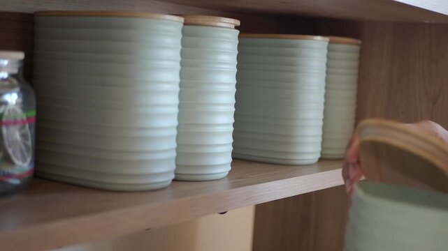 Woman arranging elegant ceramic containers with wooden lids on shelf, tidying up space with motion.