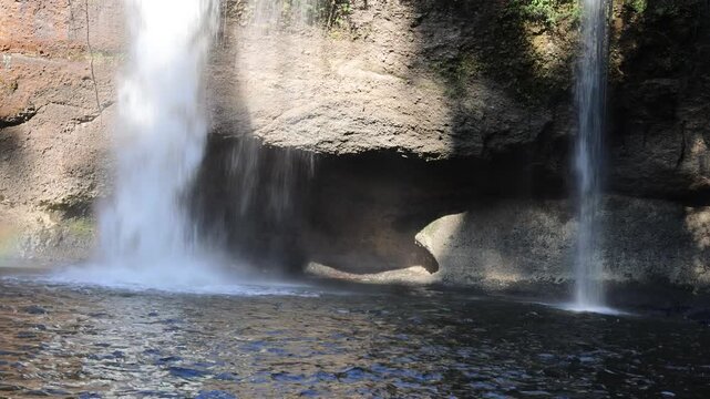 Waterfall cascading into natural rocky pool with mist and sunlight on stone cavern wall in tropical forest.