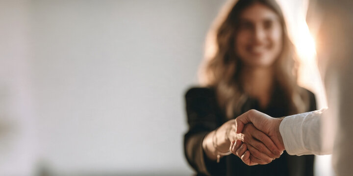 A smiling woman in a black blazer shaking hands with a business partner.