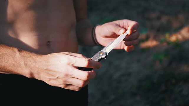 Shirtless man examines the sharp blade of a folding pocket knife outdoors. Warm afternoon sunlight highlights his hands and muscular torso against a soft forest background.