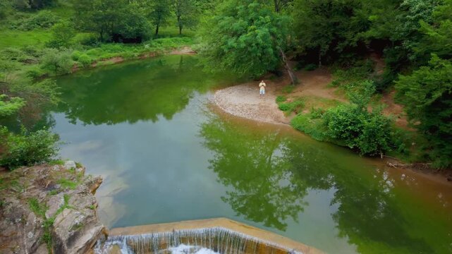 Aerial shot of drone pilot flying quadcopter on riverbank driving back to reveal stunning Samtsvera Waterfall in Georgia and a beautiful green forest landscape showcasing modern technology and nature