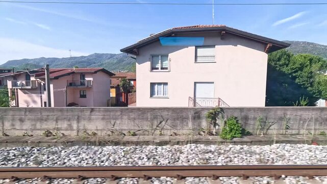 Dynamic train window view capturing houses, mountains, and a fence on a sunny day during a journey.