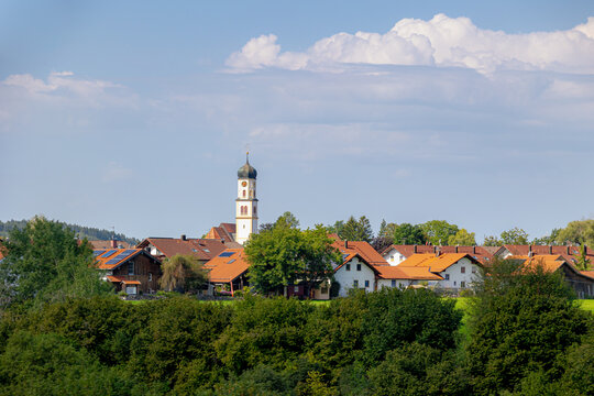 Countryside landscape, Traditional houses and church on rolling hill, Bavarian village with lush green grass meadow against majestic Alpine mountains under blue sky and white cloud in Bavaria, Germany