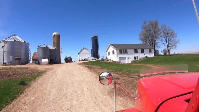 Red truck cab approaches gravel farmyard, entering past large corrugated grain silos and concrete storage tower with white barn nearby under clear blue sky