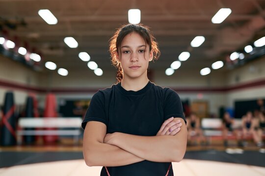 Teenage female wrestler standing confidently at center of training mat with arms crossed and focused expression, practice dummies and benches softly blurred behind