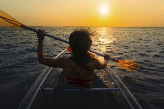 Silhouetted pretty sporty fit woman paddles clear kayak during vibrant Zanzibar sunset. Back view capturing peaceful golden hour on the Indian Ocean during a tropical tour with evening breeze