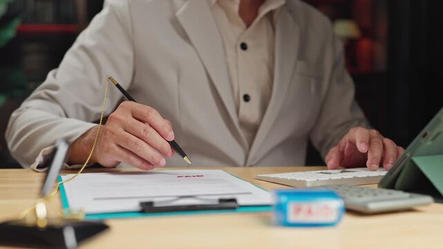 Accountant processes payment paperwork beside paid stamp, verifying each payment document before stamping final payment confirmation.