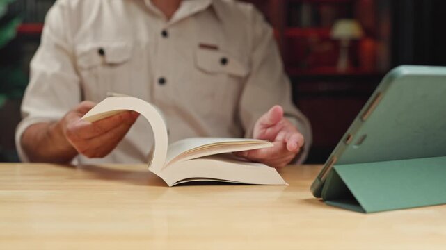 Focused adult reading quietly begins reading literature beside tablet computer, reading chapters carefully during personal study at wooden table.