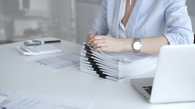 Woman's hands signing and checking financial documents, surrounded by a high stack of paperwork, a calculator, and a laptop, symbolizing busy office work, auditing, and bureaucracy. Audit and taxes