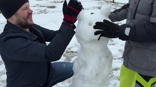 In a frosty park, a father teaches his son how to roll snowballs to build a classic snowman before their play turns into a friendly snowball fight.