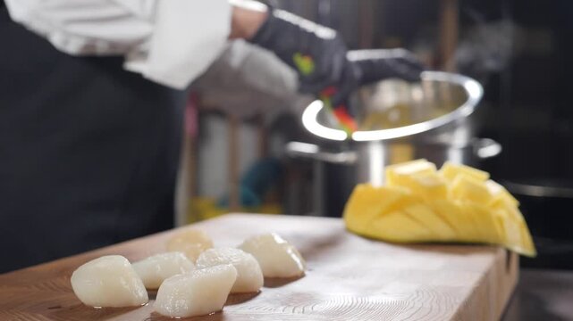 Professional chef in uniform and black gloves cooking in kitchen. Raw squid pieces and sliced yellow mango on wooden cutting board, metal pot with sauce on stove in background. Slow motion 4k video