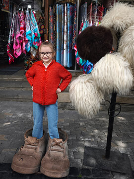 Cute young girl wearing glasses and a red jacket standing inside oversized giant shoes in front of a souvenir shop in Old City Baku, Azerbaijan. Surrounded by traditional Caucasian papakha fur hats 