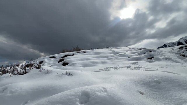 A dramatic wide view of a snow-covered alpine field and hillside under dark, heavy storm clouds with sunlight breaking through the overcast sky in Khyber Pakhtunkhwa, Pakistan,