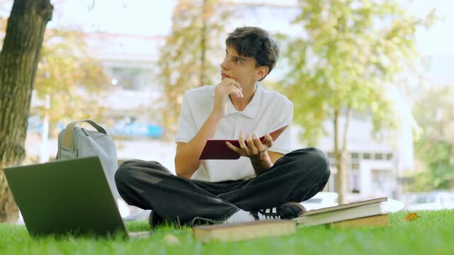 Attentive male learner looking away while writing school tasks on the grass.