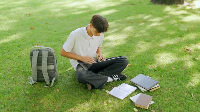 Caucasian teenage scholar working on a computer on the outdoor campus lawn.