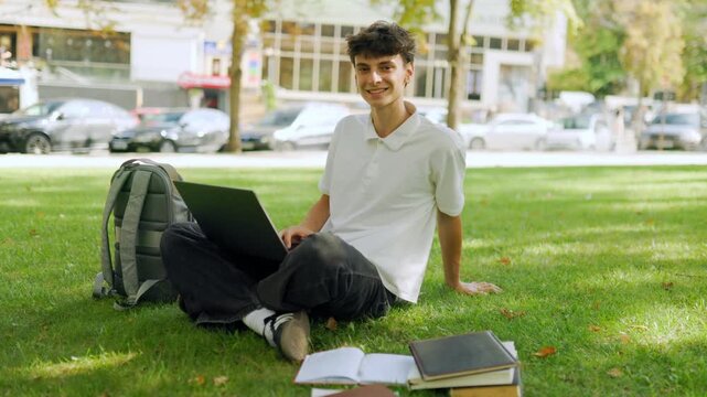 Smiling male learner with a laptop looking at the camera on the park grass.