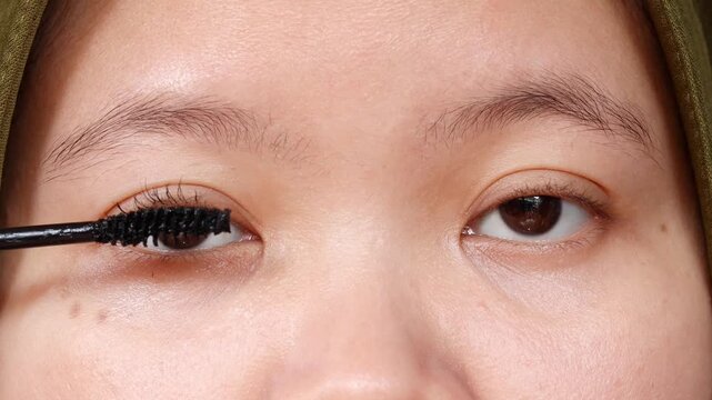 Asian Muslim woman applying black mascara to her eyelashes as part of a daily beauty and makeup routine at home.