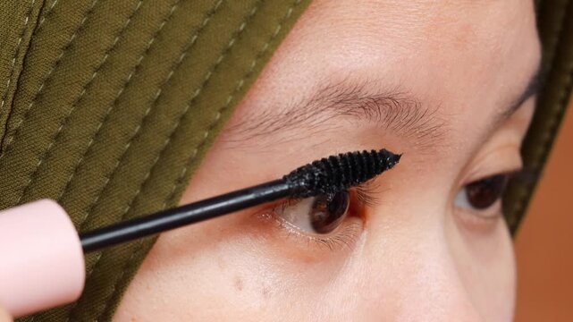 Asian Muslim woman applying black mascara to her eyelashes as part of a daily beauty and makeup routine at home.