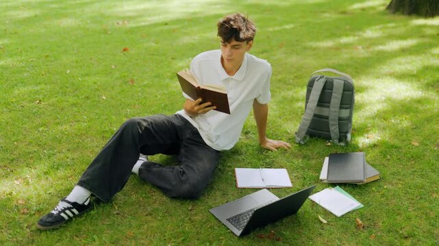 Focused young student reading an educational book on the campus grass.