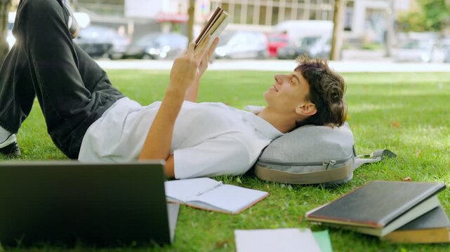 Relaxed male learner reading a book while lying on the green campus lawn.