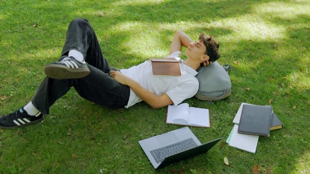 Relaxed young guy resting with a book on his chest on the park grass.