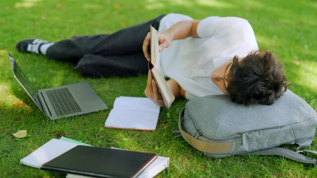 Relaxed teenage learner resting with a book on the campus grass.