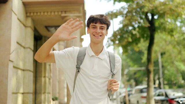 Smiling teenage scholar waving his hand directly at the camera outdoors.
