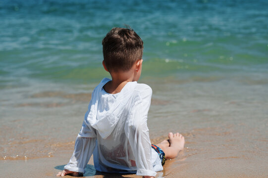 A boy is sitting on the sand on the shoreline of the sea.