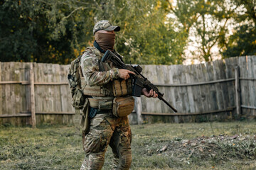 Fototapeta premium Military soldier posing at camera with assault rifle against the old wooden fence.