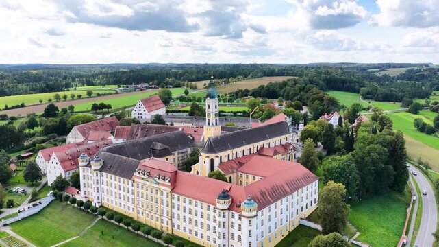 Malerische Luftaufnahme von Ochsenhausen mit der barocken Klosteranlage, eingebettet in die idyllische, gr&uuml;ne H&uuml;gellandschaft Oberschwabens unter blauem Sommerhimmel mit Wolken.