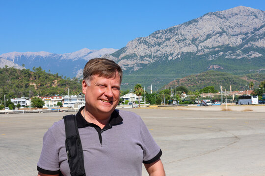 A portrait adult man squinting in the bright sun against a backdrop of blurred, majestic mountain peaks.