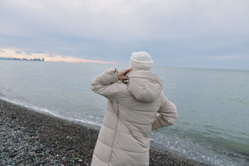 Beach seascape in winter: a person in a coat and hat stands on pebbles, gazing at the calm water and distant horizon, creating a quiet mood. © SHOTPRIME STUDIO