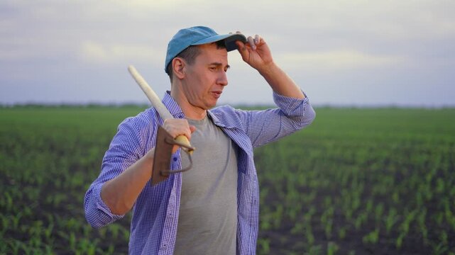 Confident farmer stands in green field holding hoe on shoulder. Man in cap talks and gestures with hand. Farmer works in agriculture field with young crops. Farmer looks over growing plants under sky.