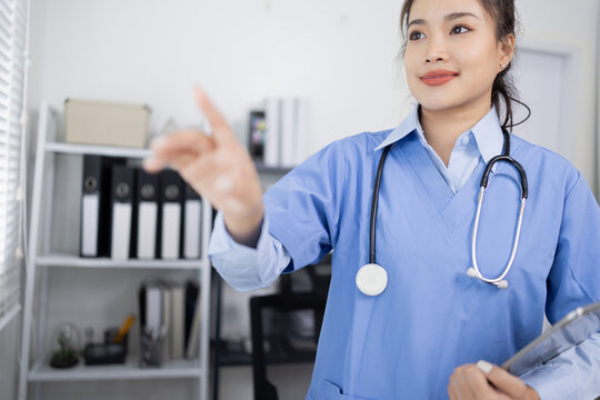 Asian female doctor in blue scrubs, wearing a stethoscope, holds a tablet and points to the right side of the frame in a modern clinic office.