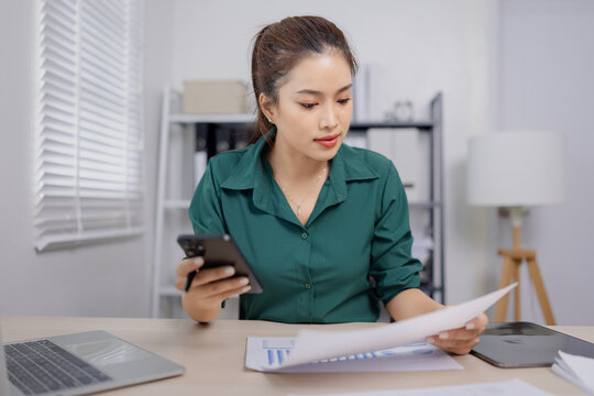 Asian businesswoman working in an office, reviewing documents while using a smartphone.